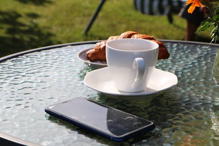 Woman in the summer has breakfast in a summer cafe with a cup of coffee, croissant and a smartphone