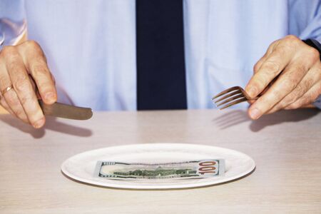 Close-up - money is lying on a white plate. Businessman is sitting at the table with fork and knife. White background.の写真素材
