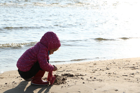 Child girl playing by the sea in autumn. A girl in a red jacket in a beautiful autumn on the seashore. Counterlight.の写真素材