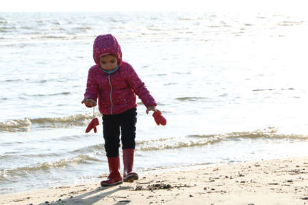 Child girl playing by the sea in autumn. A girl in a red jacket in a beautiful autumn on the seashore. Counterlight.の写真素材
