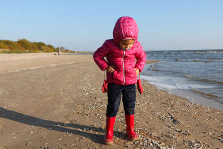 Child girl playing by the sea in autumn. A girl in a red jacket in a beautiful autumn on the seashore. Counterlight.の写真素材