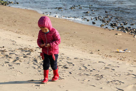 Child girl playing by the sea in autumn. A girl in a red jacket in a beautiful autumn on the seashore. Counterlight.の写真素材