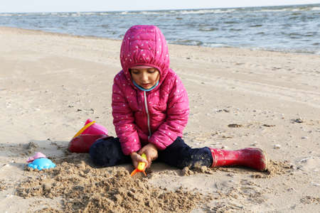Child girl playing by the sea in autumn. A girl in a red jacket in a beautiful autumn on the seashore. Counterlight.の写真素材