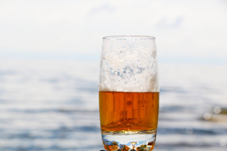 Golden foamy drink in a transparent glass on the background of the sea and the beach.. Beer with foam in a glass with a beautiful background.の写真素材