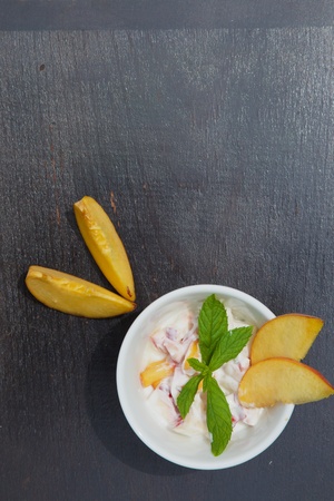 Homemade peach yogurt in a white dish on the black wooden surface. Decorated with two slices of fresh peach and fresh peppermint leaves. Top view. Free space for a textの写真素材