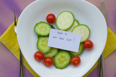 Message on the plate with raw vegetables: \"Back to raw food diet.\" Food background: fork,knife,cucumbers,cherry tomatoes,raw zucchini. The plate is place on a orange napkin on a violet surface.の写真素材