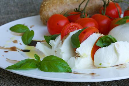 Buffalo mozzarella served with cherry tomatoes,basil leaves,and vinegar on a white plateの写真素材