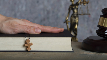 Wooden cross on a bible.Witness's hand and statue of Themis and judge's gavel in the background.の写真素材