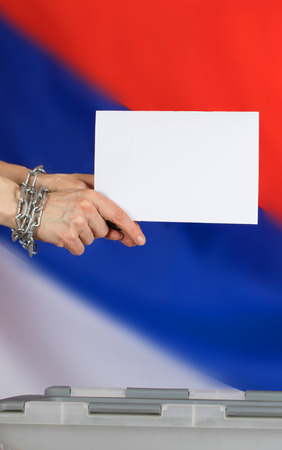 Female hands fastened by metal chain cast ballot paper in the ballot box. Russian flag in the background.の写真素材