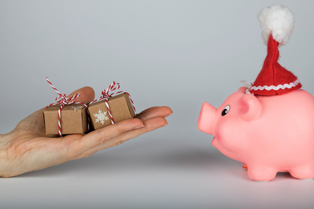 Pink piglet in a Santa Claus hat and human hand with Christmas presents in front of it. Closeupの写真素材