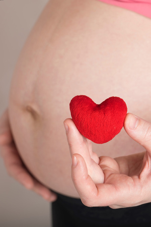 Young pregnant between 30 and 35 years old woman keeps small red plush heart close to her belly. Closeupの写真素材