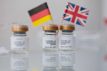 Vials with COVID-19 vaccine and syringe on a glass surface. German and British flags in the...