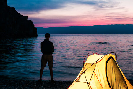 Man stand close to tent and lake shore and looking at sunsetの写真素材