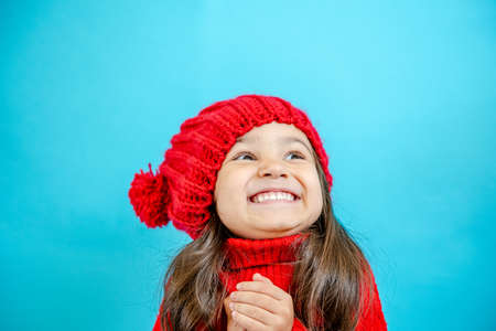 Portrait of a little curly-haired girl in a knitted red hat in winter. little girl with dark hair in a red knitted hat and sweater smiling on a blue background isolate, place for text. Winter clothesの写真素材