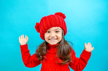 Portrait of a little curly-haired girl in a knitted red hat in winter. little girl with dark hair in a red knitted hat and sweater smiling on a blue background isolate, place for text. Winter clothesの写真素材