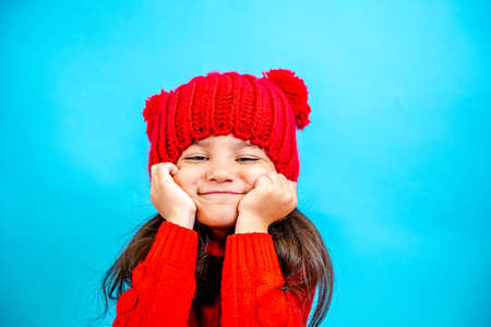 Portrait of a little curly haired girl in a knitted red hat in winter. little girl with dark hair in a red knitted hat and sweater smiling on a blue background isolate, place for text. Winter clothesの写真素材
