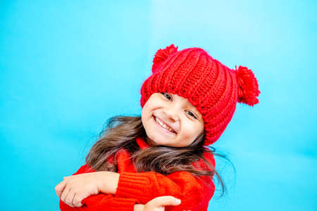 Portrait of a little curly haired girl in a knitted red hat in winter. little girl with dark hair in a red knitted hat and sweater smiling on a blue background isolate, place for text. Winter clothesの写真素材