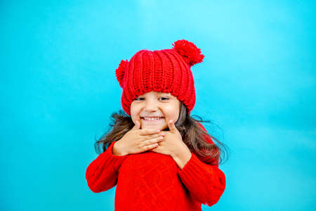 Portrait of a little curly haired girl in a knitted red hat in winter. little girl with dark hair in a red knitted hat and sweater smiling on a blue background isolate, place for text. Winter clothesの写真素材