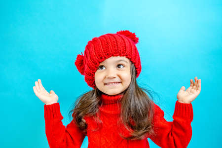 Portrait of a little curly haired girl in a knitted red hat in winter. little girl with dark hair in a red knitted hat and sweater smiling on a blue background isolate, place for text. Winter clothesの写真素材
