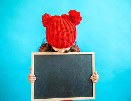Portrait of a little curly haired girl in a knitted red hat in winter. little girl with dark hair in a red knitted hat and sweater smiling on a blue background isolate, place for text. Winter clothesの写真素材