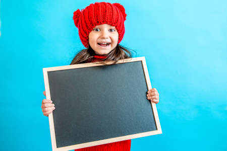 Portrait of a little curly haired girl in a knitted red hat in winter. little girl with dark hair in a red knitted hat and sweater smiling on a blue background isolate, place for text. Winter clothesの写真素材