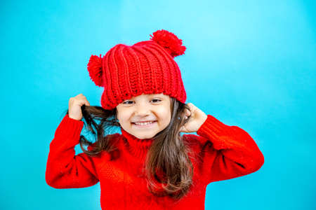 Portrait of a little curly haired girl in a knitted red hat in winter. little girl with dark hair in a red knitted hat and sweater smiling on a blue background isolate, place for text. Winter clothesの写真素材
