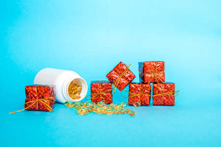 Vitamin medicinal capsules are poured out of a white bottle on a blue background, gift boxes are standing next to it. Healthy lifestyle. Pill gift for the new yearの写真素材