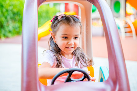 Cheerful little girl with two ponytails sits in a children's car on the playground in the parkの写真素材