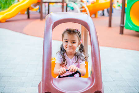 Cheerful little girl with two ponytails sits in a children's car on the playground in the parkの写真素材
