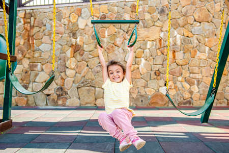 A little cheerful girl plays on vacation in the park on a children's playground. The child is engaged on gymnastic rings on the street on vacation. The child goes in for sports on simulators.の写真素材