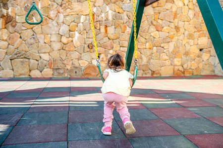 A little cheerful girl plays on vacation in the park on a children's playground. Happy child has fun on a swing in the park. Fun holidaysの写真素材