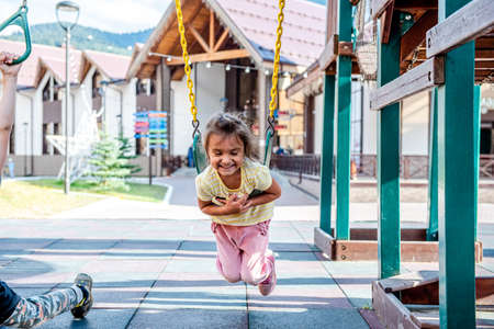 A little cheerful girl plays on vacation in the park on a children's playground. Happy child has fun on a swing in the park. Fun holidaysの写真素材