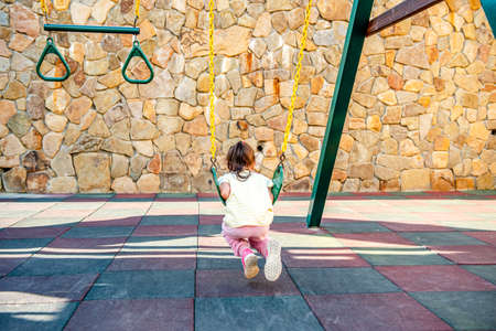A little cheerful girl plays on vacation in the park on a children's playground. Happy child has fun on a swing in the park. Fun holidaysの写真素材