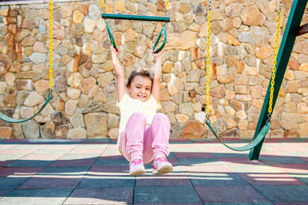 A little cheerful girl plays on vacation in the park on a children's playground. The child is engaged on gymnastic rings on the street on vacation. The child goes in for sports on simulators.の写真素材