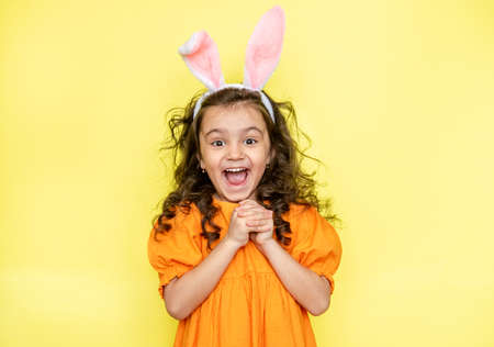 Happy curly little girl wears bunny ears, loves the celebration and mystery of the holiday, dressed in orange bright dress, isolated on yellow background. Seasonal holiday conceptの写真素材