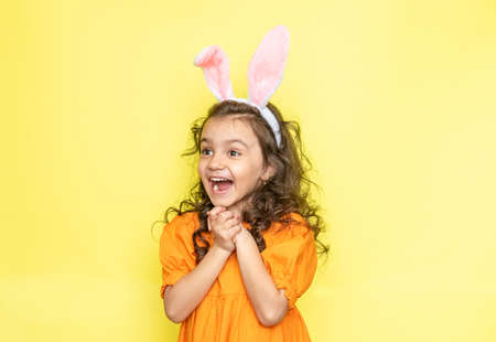 Surprised emotional curly little girl wearing bunny ears wearing bright orange dress, isolated on yellow background. Easter holiday conceptの写真素材