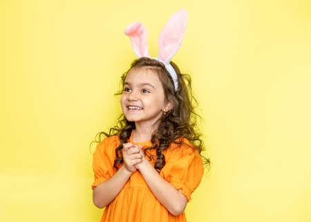 Surprised emotional curly little girl wearing bunny ears wearing bright orange dress, isolated on...