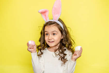 Happy curly little girl wears bunny ears raises colored Easter eggs, likes celebration and mystery...
