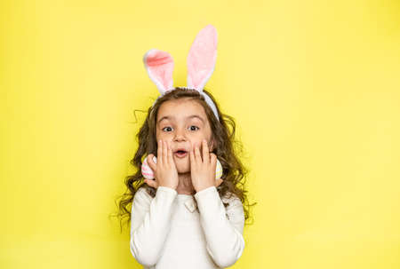 Happy curly little girl wears bunny ears raises colored Easter eggs, likes celebration and mystery of holiday, dressed in white t-shirt, isolated on yellow background. Seasonal holiday conceptの写真素材
