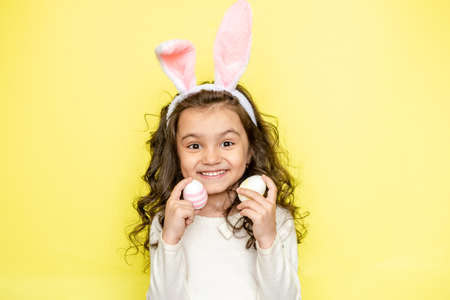 Happy curly little girl wears bunny ears raises colored Easter eggs, likes celebration and mystery of holiday, dressed in white t-shirt, isolated on yellow background. Seasonal holiday conceptの写真素材