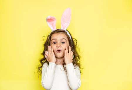 Happy curly little girl wears bunny ears raises colored Easter eggs, likes celebration and mystery of holiday, dressed in white t-shirt, isolated on yellow background. Seasonal holiday conceptの写真素材