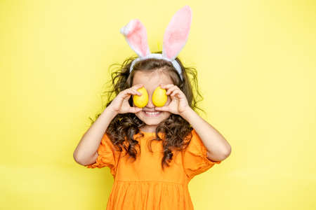 Cheerful little ethnic boy with Afro hair in Easter costume and bunny ears smiling while covering eye with colorful egg against blue backgroundの写真素材