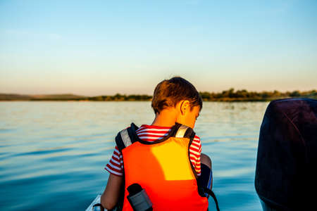 Happy boy 8 years old in a life jacket while walking on a boat. View from the back. Fun holidays on the lakeの写真素材