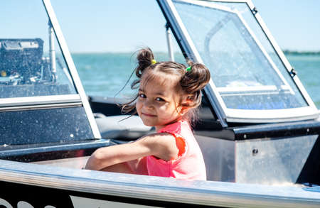 Happy little girl sits in a motor boat on the shore of a lake or river. Summer vacation. Rest with children by the water.の写真素材