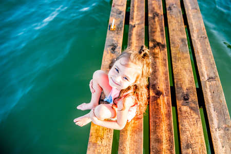 Cute little girl on the beach. The kid is resting by the sea. Summer vacation. Girl in the summer. A beautiful little girl sits on the pier by the water. girl lying on a wooden bench on the beach.の写真素材