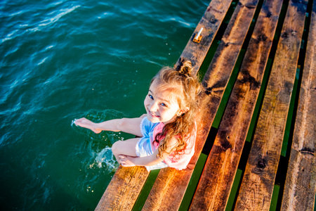 Cute little girl on the beach. The kid is resting by the sea. Summer vacation. Girl in the summer. A beautiful little girl sits on the pier by the water. girl lying on a wooden bench on the beach.の写真素材