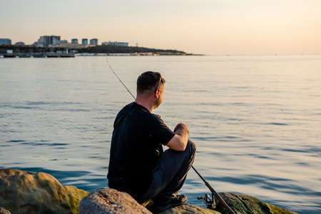 A fisherman sits on a rock by the sea with a fishing rod at sunset. Single sea fishing. Day at the beach. Copy space for textの写真素材