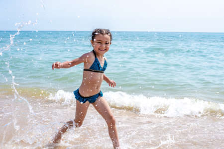 Happy little girl in a bathing suit running with open arms on the golden sand, looking at the camera, spending free time on the beach by the sea. Full length, front view. Childhood conceptの写真素材