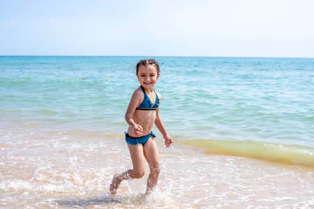 Happy little girl in a bathing suit running with open arms on the golden sand, looking at the camera, spending free time on the beach by the sea. Full length, front view. Childhood conceptの写真素材