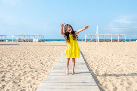 happy little curly girl in a yellow dress is jumping on the sandy beach on a sunny day. Fun holidays. Empty beachの写真素材